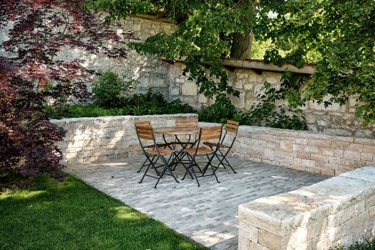 Sitzplatz mit Natursteinpflästerung, Kalksteinmauern und schattenspendenden Bäumen im Privatgarten