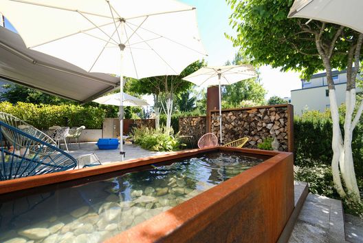 Moderner Privatgarten mit Wasserbecken aus Cortenstahl, grosszügiger Terrasse, Sonnenschirmen und üppiger Bepflanzung.