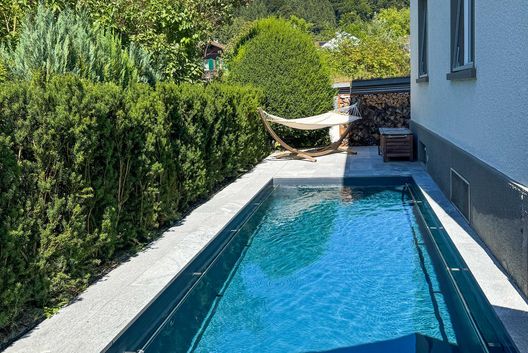 Eleganter Swimmingpool im modernen Privatgarten mit Natursteinplatten, Hängematte und Blick auf grüne Bäume und bewaldete Hügel.