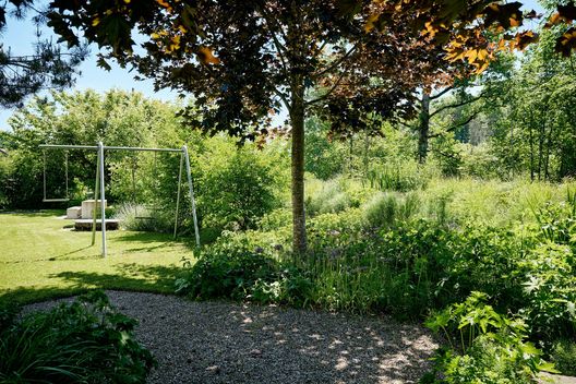 Begrünte Gartenanlage mit Kiesplatz, Schaukel und dichter Bepflanzung unter schattenspendenden Bäumen