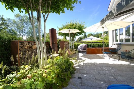 Modern gestaltete Gartenterrasse mit Natursteinbelag, Sonnenschirmen, Designer-Gartenmöbeln, Hochbeeten aus Cortenstahl und üppiger Bepflanzung in einem gepflegten Privatgarten.