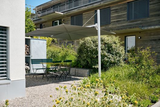 Kiesplatz mit Sonnenschirm und Sitzgruppe in einem begrünten Garten bei modernem Wohnhaus