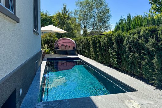 Moderner Gartenpool neben einem Einfamilienhaus mit gepflegter Hecke, Sonnenschirm und gemütlicher Outdoor-Lounge an einem sonnigen Sommertag.