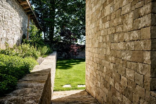 Schmaler Gartenweg zwischen Kalksteinfassade, Natursteinmauer und bepflanztem Beet